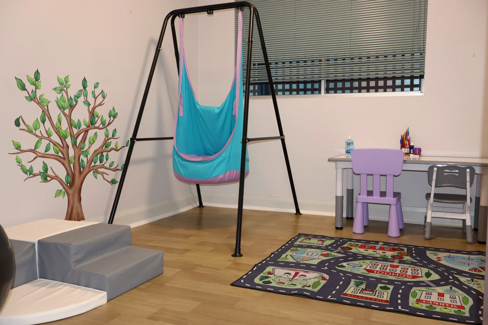 Speech therapy room at Square One with a lycra swing, child-height desk, tree wall mural and soft play steps